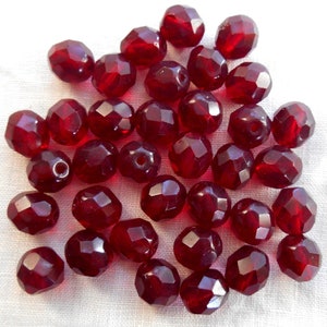 Può includere: Una collezione di circa 30 perline di vetro sfaccettate di colore rosso scuro. Le perline sono rotonde e hanno una finitura lucida.