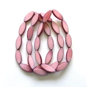 Peut inclure: Un collier composé de perles ovales rose clair. Les perles sont disposées en couches, créant un effet tridimensionnel. Les perles ont une finition brillante et sont posées sur un fond blanc.