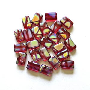 May include: A collection of rectangular glass beads in a deep red hue, each featuring an iridescent, multi-colored finish that creates a shimmering effect across the surface. The beads are arranged in a loose cluster, showcasing their smooth, polished texture and reflective quality.