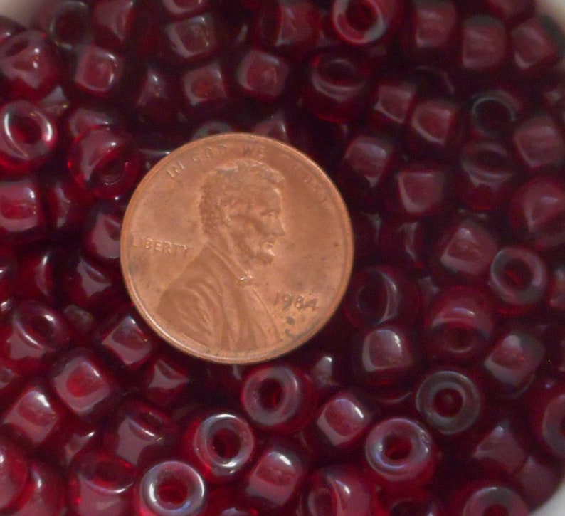 May include: A close-up image of a large quantity of small, dark red, glass beads. A US penny is included for scale.