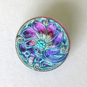 Peut inclure: Un bouton rond avec un motif floral. Le bouton présente une fleur centrale turquoise entourée de pétales violets et turquoise. Il a un bord doré et un bord rose.