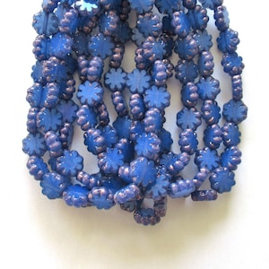 May include: Close-up of a collection of blue flower-shaped beads. The beads have a matte finish and are strung together. The edges of the beads have a darker, purplish hue. The beads are arranged in multiple strands.