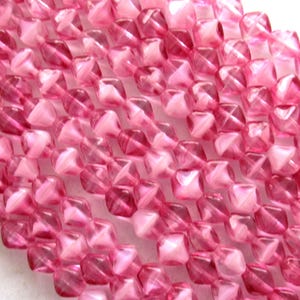 May include: A strand of pink faceted glass beads. The beads are square shaped with a slight diamond cut. The beads are translucent and have a slight iridescent sheen.