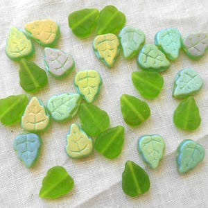 May include: A collection of leaf-shaped glass beads in various shades of green, blue, and yellow. The beads have detailed vein patterns and are scattered on a white fabric surface. These decorative beads are ideal for crafting and jewelry making.