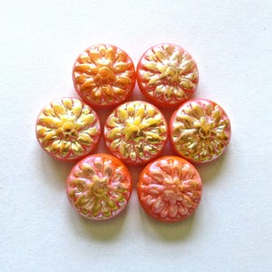 Peut inclure: Sept perles en verre rondes de couleur corail disposées en grappe. Chaque perle présente un motif floral moulé en relief sur la surface avec une finition dorée métallique appliquée sur les pétales, créant un aspect brillant et texturé.