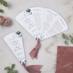 May include: A white wedding program fan with a floral design. The fan is folded and has a pink ribbon tied around it. The text on the fan reads "Welcome to the Wedding of Catherine Waters and Mark Barbare".