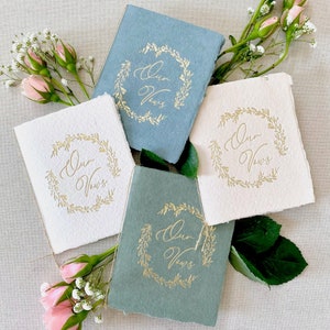 May include: Four small books with a gold foil design of a wreath with the words "Our Vows" inside. The books are on a white surface with pink and white flowers.