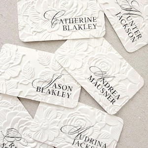 May include: White floral embossed place cards with black calligraphy names. The cards feature a detailed, raised floral design. Names include Catherine Blakley, Hunter Jackson, Jason Blakley, Andrea Mausner, Brittany Solomon, and Judrina Jackson.