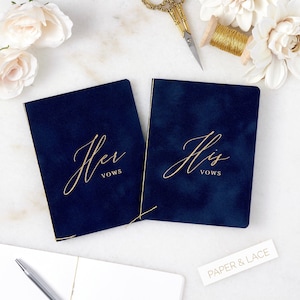 May include: Two navy blue velvet vow books with gold lettering. The book on the left says "Her Vows" and the book on the right says "His Vows".