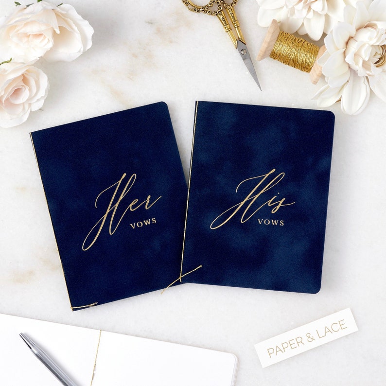May include: Two navy blue velvet vow books with gold lettering. The book on the left says "Her Vows" and the book on the right says "His Vows".