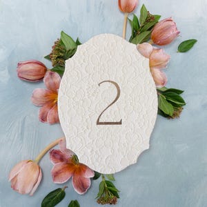 May include: An off-white, scalloped-edge card featuring a textured floral lace pattern and the number 2 printed in a metallic bronze font, surrounded by several pink and peach-colored tulips with green leaves and small brown flower clusters.