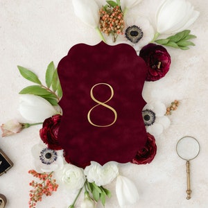 May include: A deep burgundy scalloped table number card featuring the gold numeral 8 in the center, surrounded by an arrangement of white tulips, white anemones with dark centers, deep red ranunculus, and small orange berries.