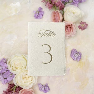 May include: A cream-colored table number card with the word "Table" and the number "3" in a gold script font. The card is surrounded by white, pink, and purple flowers on a textured, light-colored background.
