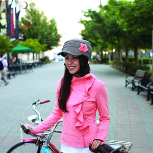 Puede incluir: Una persona lleva una chaqueta rosa con volantes en la parte delantera y un sombrero gris con una flor rosa. Está de pie junto a una bicicleta en una pasarela pavimentada con ladrillos, con árboles y bancos al fondo.