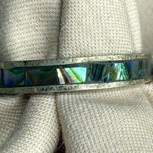 May include: A close-up of a silver ring with a band of iridescent abalone shell inlay. The shell displays shades of green, blue, and purple. The ring is set against a neutral fabric background.