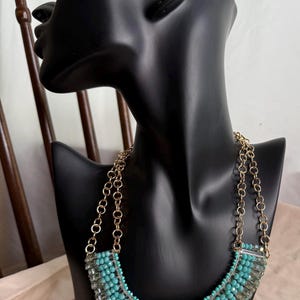 May include: A turquoise and clear beaded necklace with gold-colored chain. The necklace is displayed on a black mannequin bust. The beads are arranged in a curved pattern, creating a statement piece. The background includes a wooden chair.