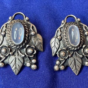 May include: A pair of silver-toned earrings with an oval, light-colored stone center. The earrings feature a leaf and floral design around the stone, with a loop at the top for wear. The earrings are set against a blue background.