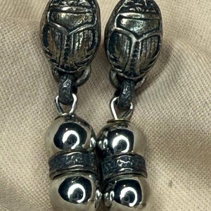 May include: A pair of silver-toned earrings. Each earring features a scarab beetle design at the top, connected to a series of rounded beads. The earrings have a vintage or antique appearance, with intricate detailing and a polished finish.