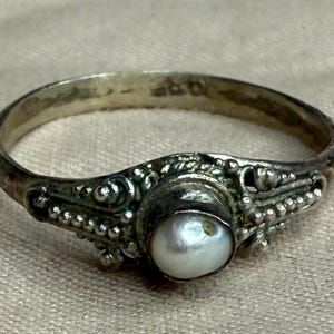 May include: A silver ring with an ornate design and a central pearl. The ring features detailed filigree work and small beaded accents. The pearl is round and has a lustrous, off-white color. The ring is likely vintage or antique.
