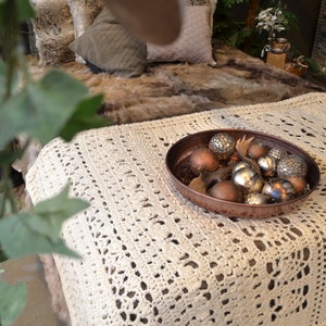 May include: A white crocheted throw blanket with a copper bowl filled with silver and brown ornaments. The bowl is sitting on top of the blanket.