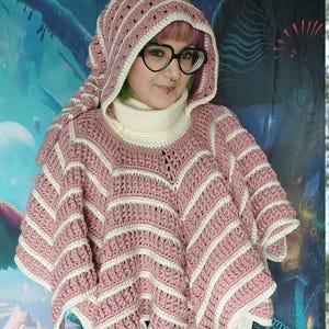Op de afbeelding: Een gehaakte poncho met capuchon voorzien van afwisselende horizontale strepen van oudroze en crèmekleurig garen. Het kledingstuk heeft een hoge crèmekleurige geribbelde kraag en een puntige capuchon. De haakstructuur is overal consistent.