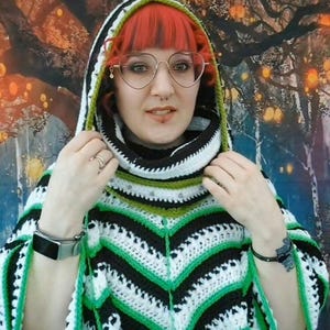 Puede incluir: Un poncho con capucha que presenta un patrón de ganchillo con rayas horizontales en blanco, negro y verde brillante. La prenda incluye un cuello alto y una capucha puntiaguda con un borde verde, confeccionada con hilo en diseño chevron.