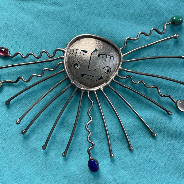 May include: A silver-toned brooch featuring a central circular face with etched eyes, nose, and mouth. Radiating from the center are multiple thin metal spokes, some straight and some wavy, tipped with small, colorful oval cabochon gemstones.