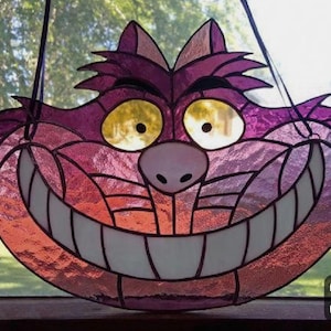 Peut inclure: Un chat du Cheshire en vitrail, avec un large sourire, dans des tons de violet, rose et orange. Le chat a des yeux jaunes et un nez rose. Des lignes noires définissent les traits. Conçu pour être suspendu, il ajoute une touche fantaisiste.