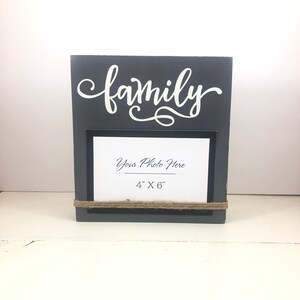 May include: Gray wooden picture frame with the word "family" in white script. The frame has a 4x6 inch opening for a photo.