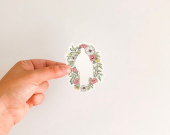 Puede incluir: Una pegatina blanca con un diseño de corona de flores. La corona está hecha de flores rosas, blancas y amarillas y hojas verdes. El centro de la corona está vacío, creando una silueta de una persona.