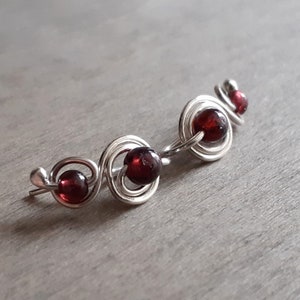Peut inclure: Boucles d'oreilles grimpantes en argent avec trois perles rondes rouge foncé. Les perles sont serties dans un design tourbillonnant en fil de fer. Les boucles d'oreilles ont une forme incurvée.