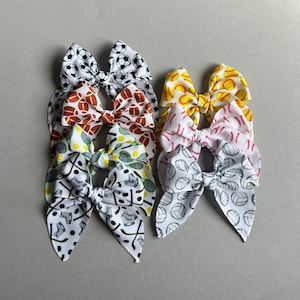 May include: Set of six hair bows with various sports-themed prints, including baseballs, basketballs, soccer balls, and tennis rackets. The bows are made of white fabric with colorful prints.