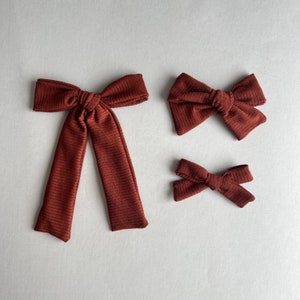 May include: Three brown fabric hair bows of different sizes. The largest bow is tied in a knot and has long tails. The other two bows are smaller and have a more traditional bow shape.