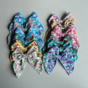 May include: A stack of 10 colorful hair bows with floral patterns. The bows are made of fabric and have a variety of colors, including pink, blue, green, yellow, and red.