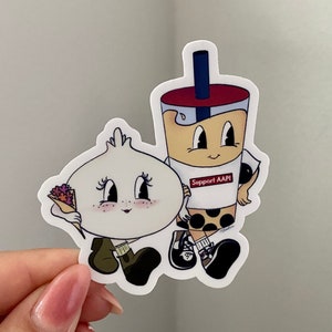 Puede incluir: Una pegatina con dos personajes de dibujos animados: un personaje blanco redondo con un ramo de flores y un personaje de té de burbujas marrón con el texto "Support AAPI".