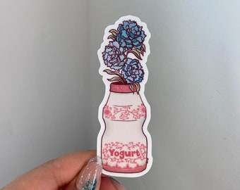 Pode incluir: Uma garrafa de iogurte branca com um design floral rosa e branco e a palavra "Iogurte" impressa na frente. Um buquê de flores azuis sai do topo da garrafa.