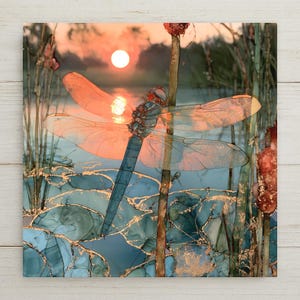May include: A square wall art print featuring a blue dragonfly with translucent wings resting on a reed. The scene depicts a sunset over water with blue and teal lily pads accented by gold leaf details, creating a reflective and layered visual effect.
