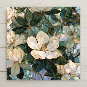 May include: A square wall art piece featuring a stained glass style illustration of white magnolia flowers with yellow centers and dark green leaves. The design includes thin gold outlines and a background of light blue and teal glass segments.