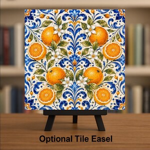 May include: A square decorative tile featuring a symmetrical pattern of bright orange citrus fruits, white blossoms, and green leaves set against a cream background with intricate blue and gold scrolling flourishes, displayed on a black easel.