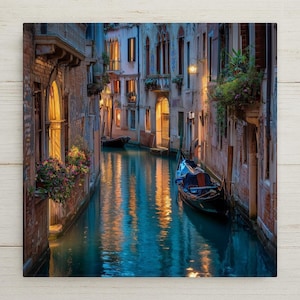 May include: A narrow canal in Venice features a dark gondola floating on blue water, reflecting the warm golden light from building windows. Stone buildings with balconies, arched doorways, and hanging flower boxes line the water's edge.