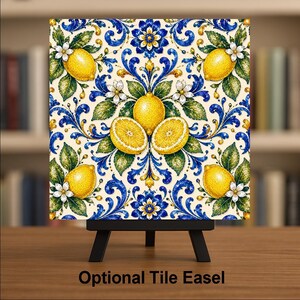 Puede incluir: Un azulejo cuadrado de cer&aacute;mica con un patr&oacute;n repetitivo de limones amarillo brillante, hojas verdes y flores blancas sobre un fondo crema con intrincados adornos en azul y dorado. El azulejo se exhibe sobre un caballete de metal negro.