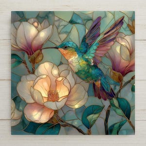 May include: A square wall art piece featuring a stained glass style illustration of a hummingbird in flight surrounded by large white and pale pink magnolia flowers. The background consists of teal and blue geometric shapes with dark outlines.