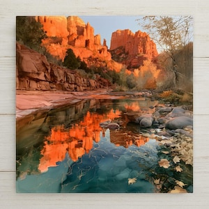 May include: A landscape photograph showing vibrant orange and red rock formations reflecting in a calm, clear stream. The water surface displays ripples and golden light, with smooth river stones and fallen autumn leaves along the shoreline.