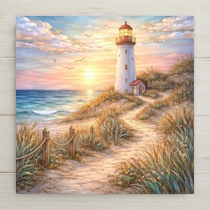 May include: A square wall art print depicting a white lighthouse standing on a grassy dune at sunset. A stone path leads toward the structure, bordered by a wooden post and rope fence. The sky is filled with soft clouds in shades of pink and gold.