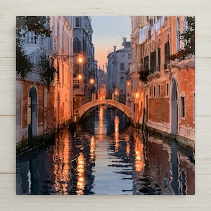 May include: A square wall art print depicting a Venetian canal at twilight. Tall, multi-story buildings with orange and beige facades line the water. A stone arched bridge spans the canal, illuminated by warm glowing lights reflecting on the water.