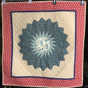May include: A handmade quilt with a blue and white starburst design in the center, surrounded by a beige background and a red and white border.