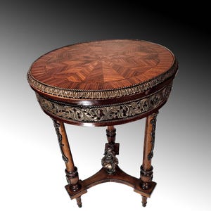 Puede incluir: Una mesa redonda de madera con una tapa de marqueter&iacute;a y patas talladas ornamentadas. La mesa tiene un borde dorado alrededor de la parte superior y un dise&ntilde;o floral decorativo en las patas.