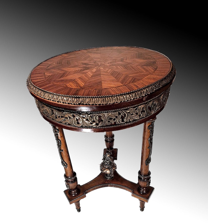 Puede incluir: Una mesa redonda de madera con una tapa de marqueter&iacute;a y patas talladas ornamentadas. La mesa tiene un borde dorado alrededor de la parte superior y un dise&ntilde;o floral decorativo en las patas.