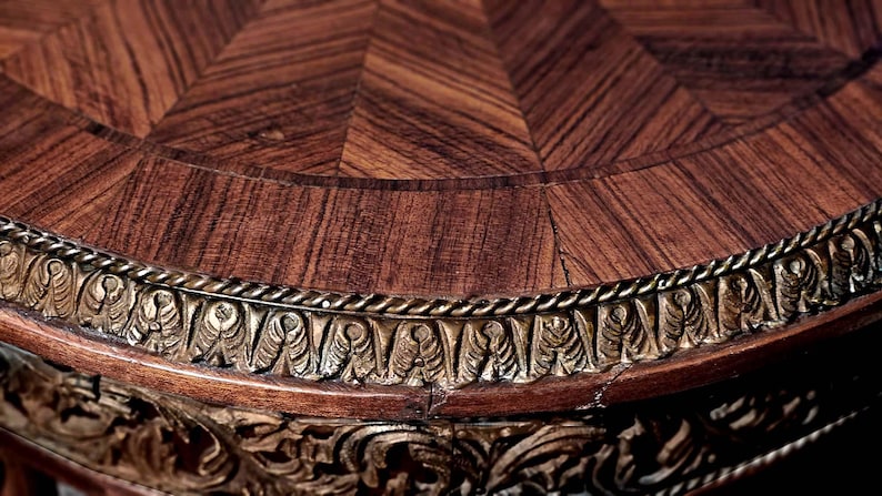 Puede incluir: Una tapa de mesa redonda de madera con un patr&oacute;n decorativo de marqueter&iacute;a y un borde dorado. La tapa de la mesa es de madera oscura con un color marr&oacute;n intenso.