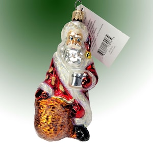 May include: A glass Christmas ornament depicting Santa Claus holding a mug and a bag of gifts. Santa is wearing a red and white suit with a white beard and a red hat. The ornament is decorated with glitter and has a gold bell on the hat.